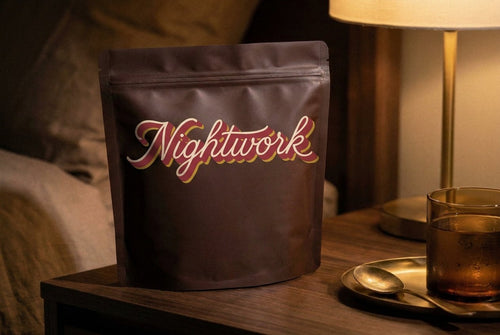 A matte chocolate Nightwork pouch on a walnut nightstand beside an amber glass and a brass spoon